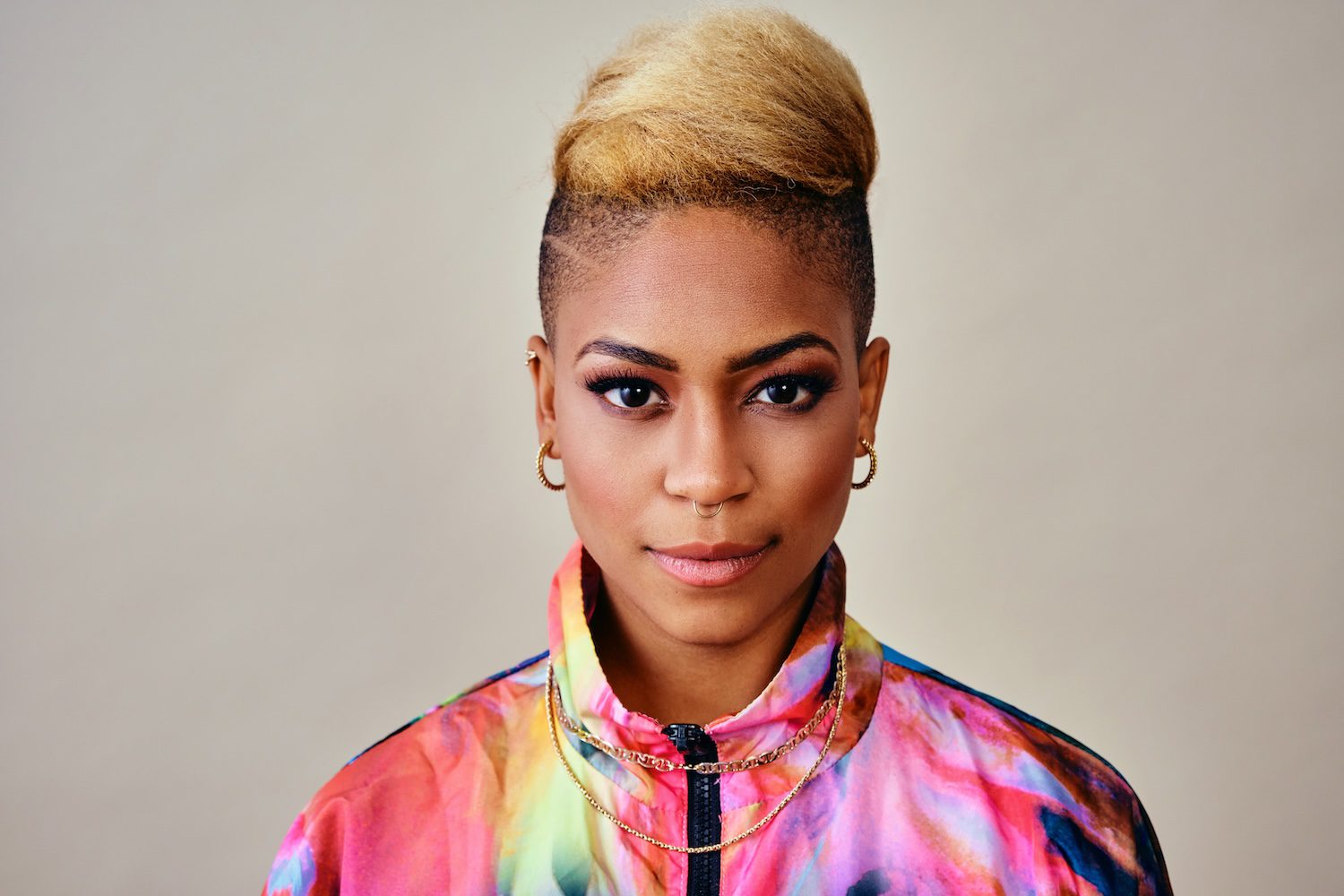 Portrait of trendy young woman with short blond hair, earrings, and colorful jacket against gray background